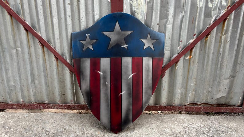 May include: A shield with a blue top section featuring three silver stars. The lower portion displays red and white stripes. The shield has a distressed, weathered appearance, suggesting a vintage or battle-worn aesthetic.