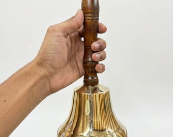 May include: A hand bell featuring a polished gold-colored brass bell base and a dark brown wooden handle. The handle has a cylindrical shape with horizontal grooves and a small brass-colored knob at the very top of the wooden grip.