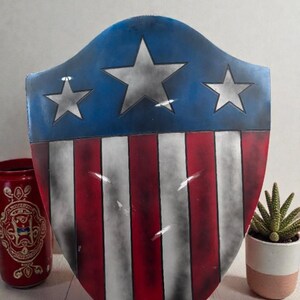 May include: A shield with a blue top section featuring three white stars. The lower section has red and white vertical stripes. The shield is a patriotic design, reminiscent of a superhero's emblem.