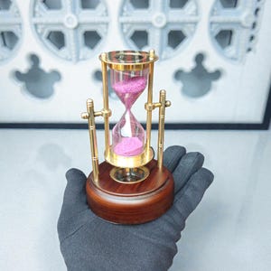 Op de afbeelding: Een zandloper met een houten basis en goudkleurige metalen accenten. De glazen kamer bevat roze zand. De zandloper wordt in een gehandschoende hand gehouden.