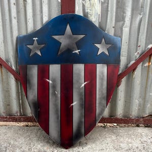 May include: A shield with a blue top section featuring three silver stars, and a red and white striped bottom. The shield has a distressed, weathered appearance, suggesting a vintage or battle-worn aesthetic.