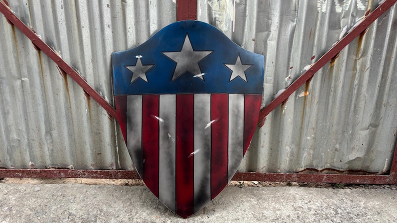 May include: A shield with a blue top section featuring three silver stars, and a red and white striped bottom. The shield has a distressed, weathered appearance, suggesting a vintage or battle-worn aesthetic.