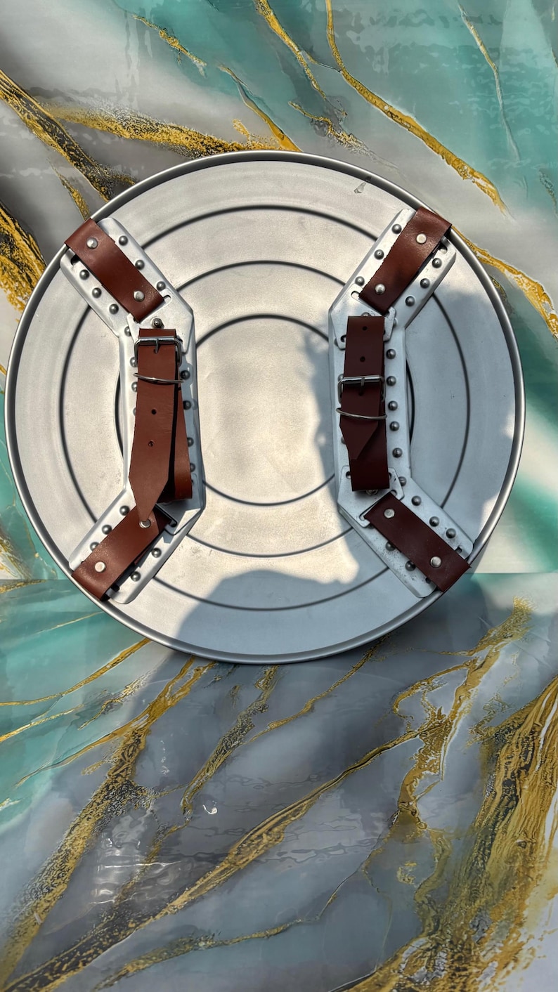 May include: A silver-colored, round shield with brown leather straps and buckles. The shield has a central circular indentation and is reinforced with metal brackets and rivets. The straps are riveted to the shield's surface.