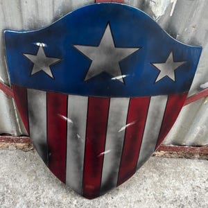 May include: A shield-shaped decorative item. The top section is blue with three grey stars, and the lower part has red and white vertical stripes. The shield has a distressed, weathered appearance.