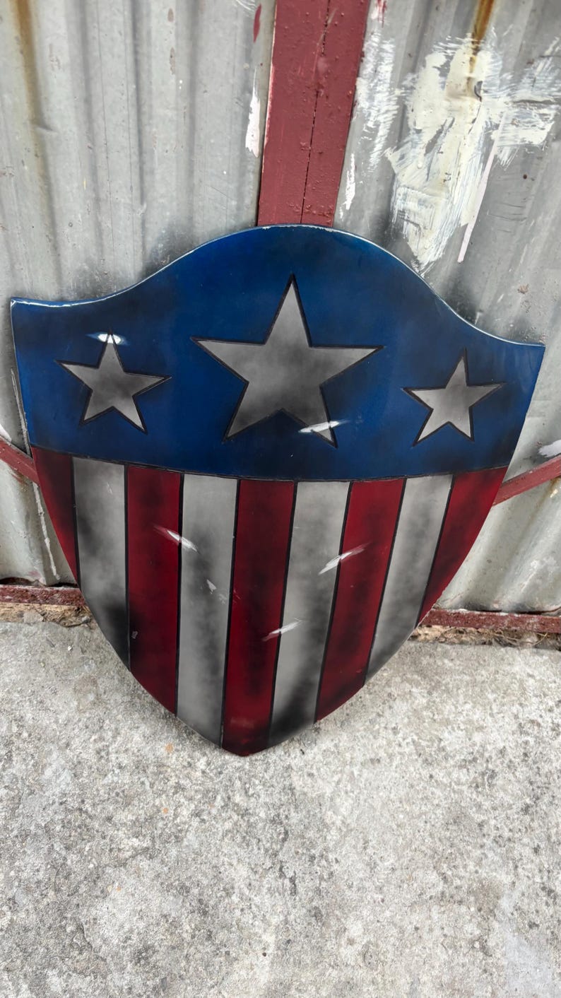 May include: A shield-shaped decorative item. The top section is blue with three grey stars, and the lower part has red and white vertical stripes. The shield has a distressed, weathered appearance.