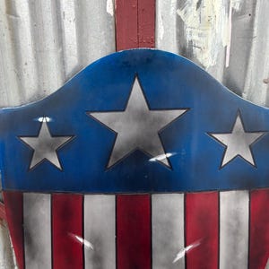 May include: A shield design in patriotic colours, with a blue top bearing three silver stars, and a red and white striped base. The shield has a distressed, vintage appearance. The background is a corrugated metal surface.