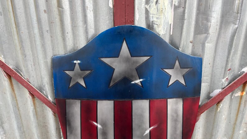 May include: A shield design in patriotic colours, with a blue top bearing three silver stars, and a red and white striped base. The shield has a distressed, vintage appearance. The background is a corrugated metal surface.