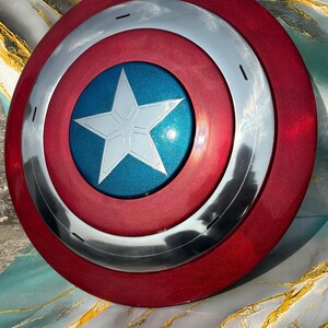 May include: A red, silver, and blue shield with a star emblem. The shield has a circular design with a red outer ring, a silver middle ring, and a blue center with a white star. The shield is a prop or collectible.