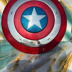 May include: A red, white, and blue shield with a star in the center. The shield has a circular design with a red outer ring, a silver middle ring, and a blue center with a white star. A superhero-themed item.