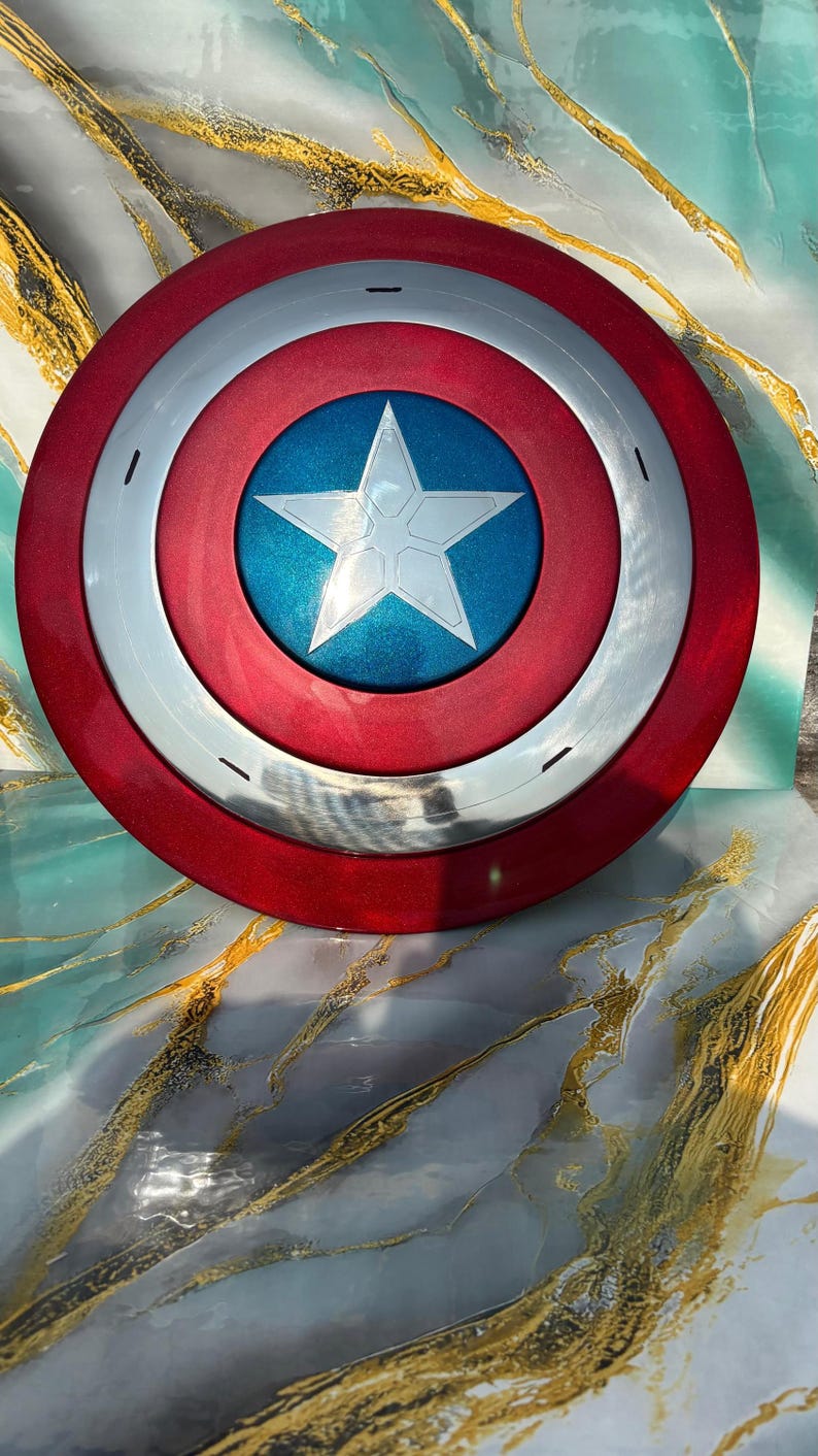 May include: A red, white, and blue shield with a star in the center. The shield has a circular design with a red outer ring, a silver middle ring, and a blue center with a white star. A superhero-themed item.