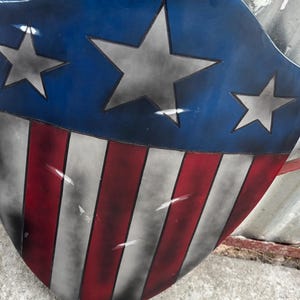 May include: A shield with a blue top section featuring three grey stars. The lower section has red and white stripes. The shield has a distressed, weathered appearance, with visible paint imperfections.
