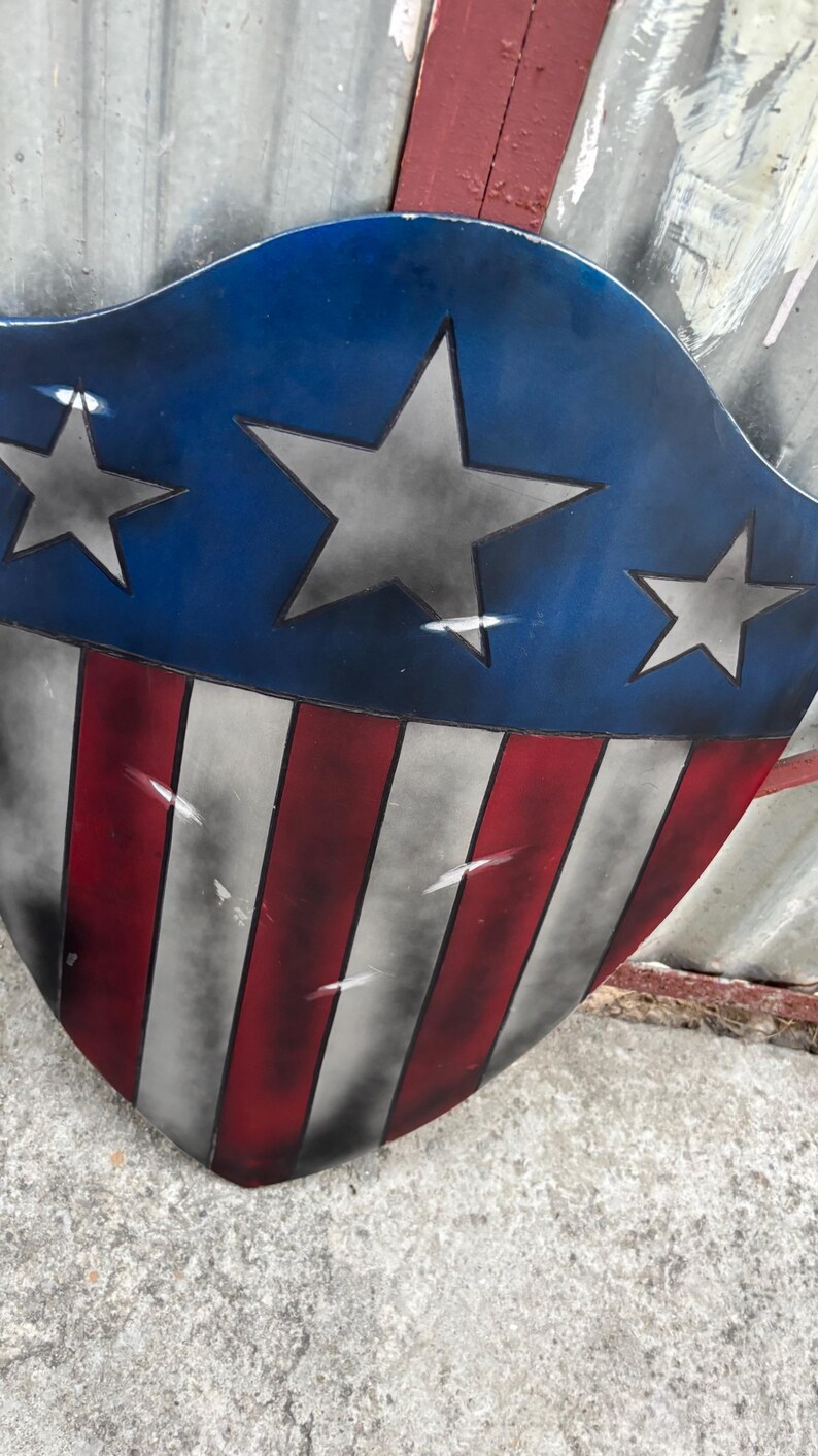 May include: A shield with a blue top section featuring three grey stars. The lower section has red and white stripes. The shield has a distressed, weathered appearance, with visible paint imperfections.