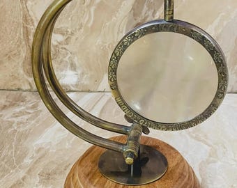 May include: A brass magnifying glass mounted on a curved brass arm and a circular wooden base. The circular frame features engraved floral patterns and the text "1941" and "HENRY HUGHES SON LTD LONDON" around the glass lens.