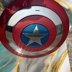 May include: A red, white, and blue shield with a silver star in the center. The shield has a metallic finish and a circular design, reminiscent of a superhero's emblem.
