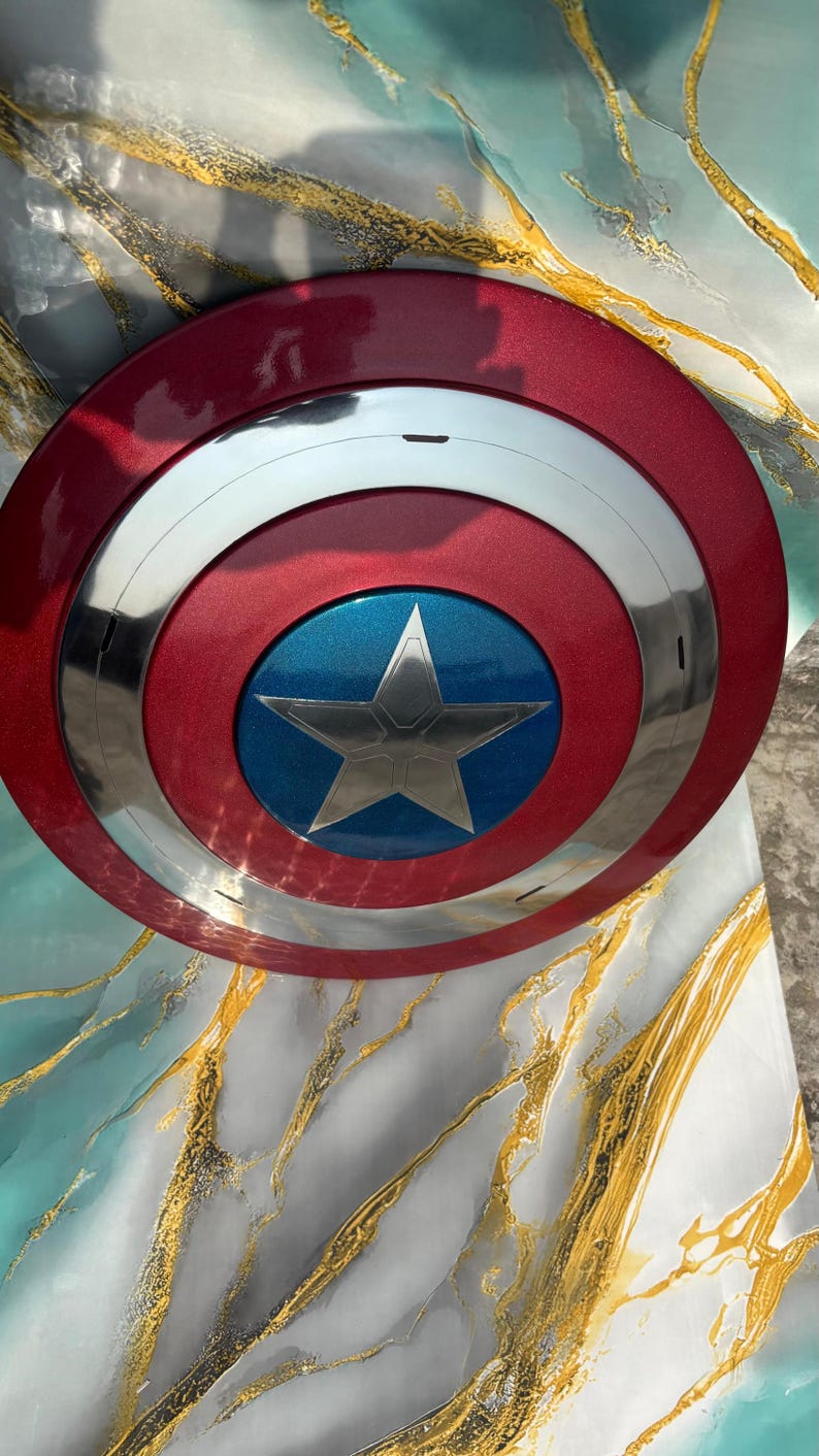 May include: A red, white, and blue shield with a silver star in the center. The shield has a metallic finish and a circular design, reminiscent of a superhero's emblem.