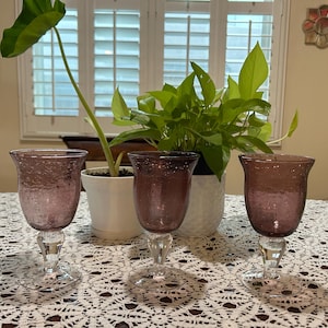May include: Three stemmed wine glasses with a textured, amethyst-colored bowl and a clear, faceted stem. The glasses are arranged on a white lace tablecloth, with green plants and a window in the background.