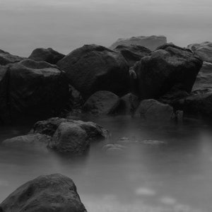 May include: A black and white photo of a rocky shoreline with fog rolling in. The rocks are large and jagged, and the water is calm and still.