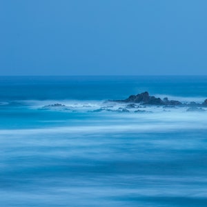 May include: A long exposure photograph of a rocky shoreline with the ocean in the background. The water is a light blue color and the rocks are a dark gray color. The water is blurred due to the long exposure.