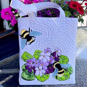 Op de afbeelding: Een witte gewatteerde tote bag met een ontwerp van paarse bloemen en twee hommels. De tas heeft witte stippen en een golvend patroon.