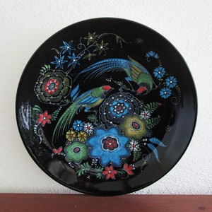 May include: A black decorative plate featuring a vibrant floral and bird design. The plate showcases two birds with blue and green plumage, surrounded by colorful flowers in shades of blue, red, and green. The plate has a glossy finish.