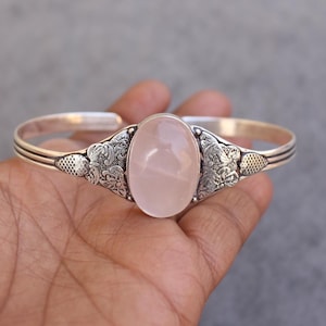 May include: A silver cuff bracelet featuring an oval, light pink rose quartz stone. The bracelet has ornate floral designs on either side of the stone and a textured pattern near the ends. The bracelet is a jewelry item.