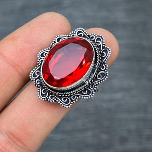 May include: A silver ring with an ornate design and a large, oval-shaped red gemstone.