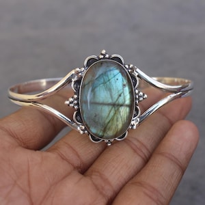 May include: A silver cuff bracelet featuring a large, oval-shaped labradorite gemstone with blue and green iridescence. The stone is set in a decorative silver frame with small bead accents, connected to a smooth, split-shank silver band.