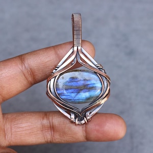 May include: A handcrafted pendant featuring an oval labradorite gemstone with blue and purple iridescence. The stone is set in a copper and silver wire wrap design, with a loop for a chain. The pendant is held in a hand.