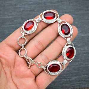 May include: A silver bracelet with five red oval-shaped gemstones. The bracelet has a lobster claw clasp.