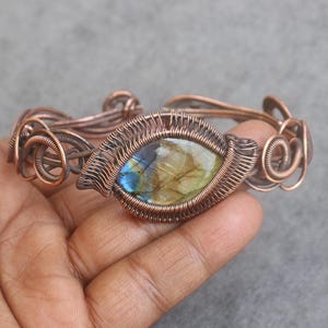 Könnte beinhalten: Kupferdraht-Armband mit einem ovalen Edelstein in der Mitte, der blaue und gelbe Farbtöne aufweist. Das Armband hat aufwendige Drahtdetails und ein handgefertigtes Aussehen. Ein einzigartiges Schmuckstück.