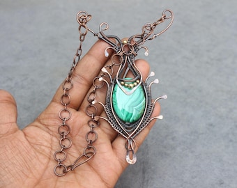 May include: A handcrafted copper wire necklace featuring a large, oval malachite stone pendant. The pendant is intricately wired with decorative elements and a chain. The necklace is held in a hand.
