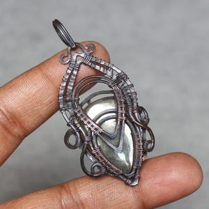 May include: A handcrafted pendant featuring a polished, iridescent stone encased in intricate wirework. The pendant's frame is made of dark, oxidized metal, with decorative swirls and loops. The stone has a metallic sheen, and the overall design is artistic and unique.