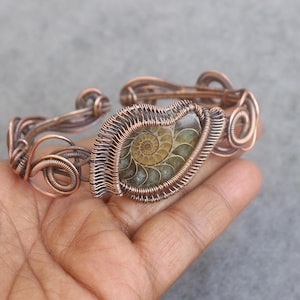 Op de afbeelding: Koperdraad gewikkelde armband met een gefossiliseerde ammoniet middensteen. De armband heeft ingewikkeld draadwerk met spiraal- en lusontwerpen. De ammoniet toont een gedetailleerd spiraalpatroon in tinten bruin en beige.