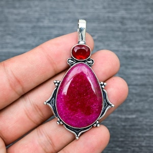 Peut inclure: Un pendentif en argent avec une grande pierre précieuse rose et une plus petite pierre précieuse rouge. Le pendentif a un design orné avec des détails complexes.
