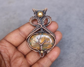 May include: A handcrafted pendant featuring a wire-wrapped design. The pendant showcases a polished, oval-shaped stone with a tree of life motif. The wirework is copper-colored, with a gold-colored accent bead. The stone has brown, green, and yellow hues.