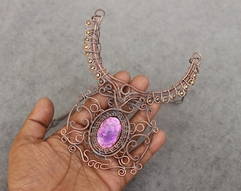 May include: A handcrafted copper necklace with a large oval purple gemstone centerpiece. The necklace features intricate wirework and decorative accents, creating a unique and artistic design. The necklace is held in a hand against a gray background.