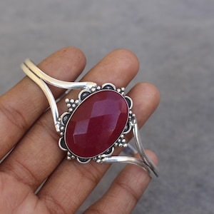 May include: A silver cuff bracelet featuring a large, oval-shaped, faceted deep red gemstone at the center. The metal setting around the stone is decorated with small silver beads and intricate wirework, while the band splits into two arms.