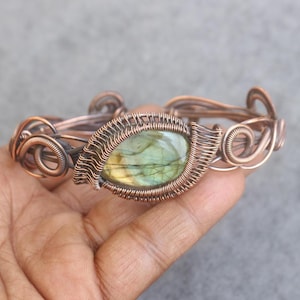 Puede incluir: Pulsera de alambre de cobre con una piedra preciosa central ovalada verde y amarilla. La pulsera tiene un intrincado trabajo de alambre con remolinos decorativos y una estética natural y terrosa.