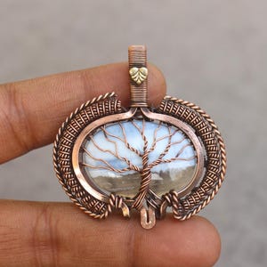 May include: A handcrafted copper pendant featuring a tree design. The pendant has a central oval-shaped stone with a landscape scene, framed by intricate wirework. A small gold-colored leaf detail is at the top.