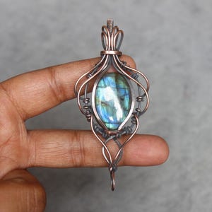 Könnte beinhalten: Handgefertigter Anhänger mit einem blau-grünen Labradorit-Edelstein, kunstvoll in Kupferdraht gewickelt. Der Anhänger hat ein verziertes, offenes Design. Der Edelstein zeigt einen schimmernden, irisierenden Effekt. Einzigartiger Schmuck.