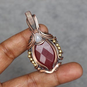 May include: A red gemstone pendant with a copper wire wrap and gold beads. The pendant is held by a finger.