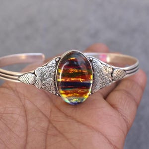 May include: Silver cuff bracelet with an oval, dichroic glass centerpiece. The glass displays a vibrant array of colors including red, orange, yellow, and blue. The bracelet features floral and textured details on the sides.