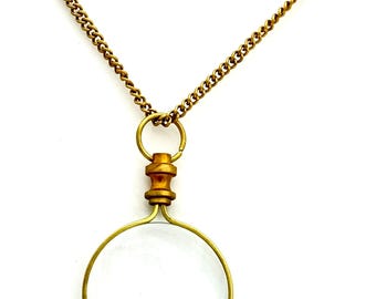 May include: A gold chain necklace with a round magnifying glass pendant. The pendant has a brass frame and a clear glass lens.