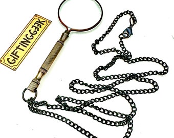May include: A vintage-style magnifying glass with a bronze-colored frame and handle, attached to a dark chain necklace. The handle has a detailed, textured design. A small rectangular sign with the text "GIFTINGGEEK" is also present.
