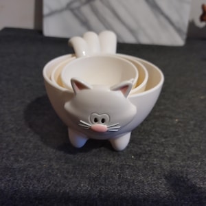 May include: A set of four white cat-shaped measuring cups. The largest cup forms the cat's body, with the smaller cups nested inside. The cat face has pink accents for the nose and ears, and white whiskers. The tail is also white.