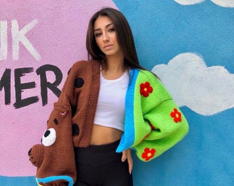 May include: A brown and green knit cardigan with a cartoon dog design on one side and flower accents on the other. The cardigan has a blue trim and black polka dots. The background features a pink and blue mural.