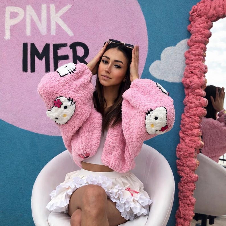 May include: A pink, fluffy jacket featuring a Hello Kitty design on the sleeves. The jacket is paired with a white crop top and a ruffled skirt. The background includes the text "PNK IMER" on a pink and blue wall.