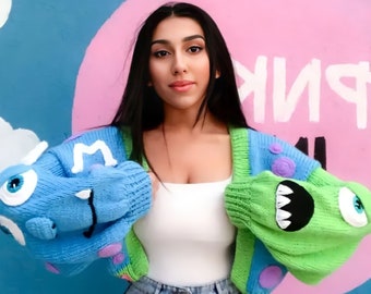 May include: A blue and green monster-themed cardigan with a white tank top. The cardigan features large, cartoonish monster arms with eyes, teeth, and purple accents. The background is a blue wall with a pink circle.