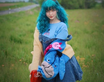 May include: A person wearing a light blue top with an Eeyore graphic, denim overalls, and a yellow sweater. They are holding a blue Eeyore hat and a red cup. The person has teal hair and is standing in a grassy field.