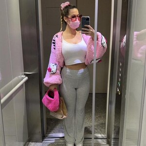 May include: A pink, fuzzy cardigan with Hello Kitty designs, paired with a white top and gray sweatpants. The outfit includes a matching pink purse and scrunchie. The person is wearing pink sunglasses and a face mask.